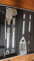 Open metal case showing BBQ utensils including tongs, spatula, fork, brush, and scraper all secured inside.
