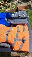 Two youth life jackets, orange with grey straps and blue with grey straps, laid out on grass with a cardboard box behind them.