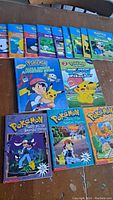 Top-down photo showing the front covers of 14 Pokemon-themed children's books arranged on a wooden surface. Books include brightly colored covers with Pikachu, Ash, and other characters from the Pokemon series.