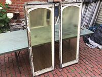 Pair of vintage decorative window sashes standing side by side outdoors on brick patio with green table behind.