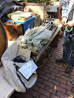 Camping bed cot folded with canvas fabric and black reinforced corners with metal rings, placed on a wooden bench surrounded by various items, including parts and instruction sheet.