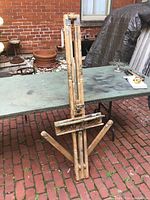 Wooden artist easel folded and resting against a table outdoors on a red brick surface. Shows adjustable arms and wooden slats with paint wear.
