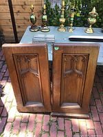 Two wooden Gothic style pew ends standing side by side outdoors on brick paving, with brass candlesticks and other metal items placed on a nearby table behind them.