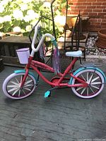 Photo of a Journey Girls toy bike in a garden setting showing side view of the bicycle with red frame, white and turquoise parts, basket, and tassels.