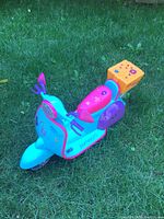 Image of a turquoise plastic ride-on toy scooter with pink seat, orange storage box with star decoration, and purple side panels, set on grass.