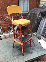 Full view of vintage kitchen step stool chair with yellow wooden seat and backrest, red metal frame, and two black metal steps.