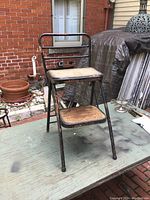 Front view of a black metal kitchen step stool with two wooden steps, the top step with a cushion pad and a small metal backrest, showing wear and rust.