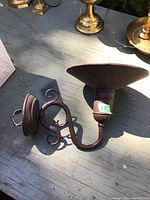 Single contemporary metal wall lamp sconce with curled arm and large conical lampshade holder, mounted on a wooden surface outdoors with sunlight casting shadows.