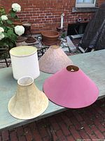 Four lampshades on outdoor table showing variety in size, shape, and color including white, beige, yellowish, and pink shades.