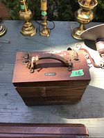 Closed wooden box with leather handle and brass hardware, placed among other brass items on a grey surface.