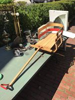 Photo of the wooden sled outside, showing the full length including handle and metal runners, with background items like lamps and garden greenery.