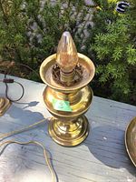 Photo shows a polished brass lamp base with an attached bulb and electrical cord placed outdoors on a wooden surface with greenery in the background.
