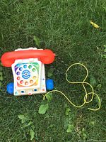 Retro Fisher Price toy telephone with red handset, white base, colorful rotary dial, and yellow cord on grass.