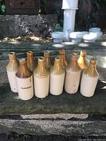 Group photo of 10 vintage UK stoneware ginger beer bottles arranged outdoors on a stone surface, showing their typical two-tone brown/beige glaze.