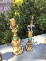 Photo showing two brass lamp bases outdoors on a white surface with greenery background. The taller lamp base has an octagonal bottom and no lampshade. The smaller lamp base has a turned finial top with an additional decorative piece near the socket.