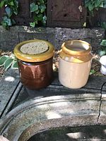 Two ceramic butter crocks placed outdoors on a stone surface. The lob and the sides show some wear consistent with use.