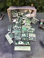 Box containing multiple modeled green fired ceramic tiles with mottled green surface glaze, placed on a wooden surface outdoors.
