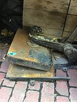 Vintage rusted desktop press with lever and rotary valve handle, on brick floor against wooden backdrop.