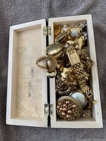 Top view of open wooden box filled with assorted gold tone vintage jewelry including bracelets, rings, brooches, and watches.