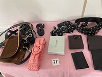Overview photo showing brown leather handbag, various belts including black studded belt, polka dot fabric belt, pink fabric belt, small brown suede pouch, and black wallets arranged on pink surface.