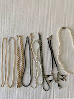 Photo of nine assorted necklaces arranged side by side on a white textured background showing various styles and materials including gold-tone chains, faux pearls, striped fabric cord, black cords with cross pendants, and cream shell or bead necklace.