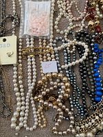 Multiple vintage necklaces arranged on a surface including faux pearl strands, gold-tone chains, and metallic bead necklaces in various colors