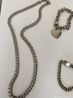 Silver chain necklace with curb links alongside two different silver bracelets, one with a heart charm and one smaller curb chain bracelet.