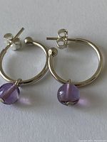 Close-up of pair of sterling silver hoop earrings with purple amethyst bead pendants shown side by side on white background.