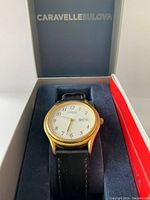 Photo showing the watch inside the open Caravelle Bulova box with visible white dial, gold-tone case, black leather strap, and red booklet.