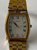 Close up front view of vintage Seiko watch face showing white dial, black Roman numerals, and gold-tone case with wear marks, with hour, minute, and second hands visible.