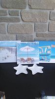Three small square beach-themed canvas prints with two white ceramic starfish dishes, placed against a brick wall and black surface.