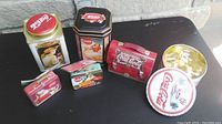 Seven Coca-Cola themed tins arranged in a row on a gray surface: two tall tins with vintage artwork, two small Christmas ornament tins, a lunchbox-style tin, a small circular tin with Christmas decorations, and a gold-colored small tin with Santa illustration.