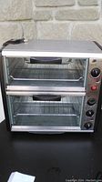 Front view of double toaster oven showing upper and lower compartments with wire racks inside, black handles on glass doors.