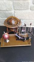 Photo showing large wood globe, small gemstone globe, black model schooner ship, and wooden tray on a black surface with light stone background.