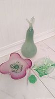 Photo of large light green art glass pear, green and purple Murano style dish, small chalet style dish, and frosted glass penguin figurine on white floor