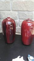 Two tall ceramic lidded decor pieces on a table in front of a stone wall. One piece is glossy deep red with black vertical stripes and the other is glossy solid red.