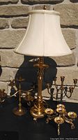 Photo of the brass table lamp with beige fabric lampshade and three brass candelabra arranged on a black surface against a stone wall background.