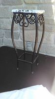Metal plant stand with black scrollwork and wood top covered by Mac tac, shown in outdoor lighting against stone wall background.