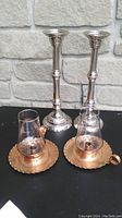 Two tall silver metal candleholders, two short copper ones with glass chimneys sitting atop them, on black surface with stone wall behind