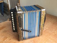 Left side angle of Hohner accordion showing detailed metal plates, blue and white bellows, and wooden knob on black key panel.