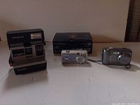 Three cameras displayed side-by-side: Polaroid Sun 600 on left, Canon PowerShot A400 center with box behind, Panasonic Lumix LS2 on right.