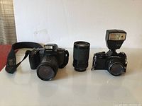 Overview of two Pentax cameras, one with external flash and 28-80mm lens, the other with built-in flash, and an additional telephoto lens between them.