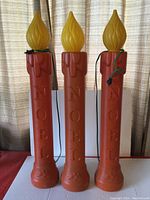 Front view showing all three orange plastic blow mold candles with yellow flame tops, each with embossed NOEL text and holly decoration near base. Two have green cords, one black cord.