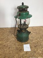 Front view of a green Coleman lantern showing glass globe and metal frame with rust.
