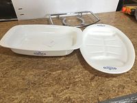 Two white Corning Ware serving dishes on a countertop, one rectangular and one divided oval plate.