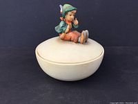 Full view of cream ceramic round covered dish with child figurine on lid, sitting pose in green and orange clothing with flute.