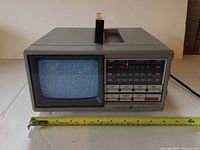 Front view of device showing screen and frequency bands with controls below and on side with measuring tape for scale.