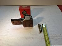Two lighters shown with one opened Jumbo lighter with a wood grain case and a small brass WWI style trench lighter next to a measuring tape.