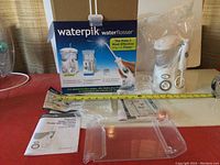Photo shows the full Waterpik Water Flosser Ultra with packaging, manual, and flosser unit with plastic seal. A measuring tape shows scale.