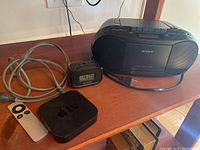 View of the Apple TV, its remote, power cord, Sony CD radio cassette-corder, and Sharp battery operated alarm clock arranged on a wood surface.